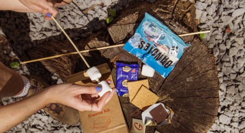 Hands preparing a s'mores kit with marshmallows, graham crackers, and chocolate on a wooden stump.