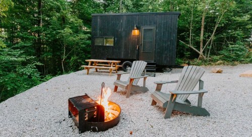 A cozy tiny house in the woods, with a fire pit and two chairs on a gravel area surrounded by trees.