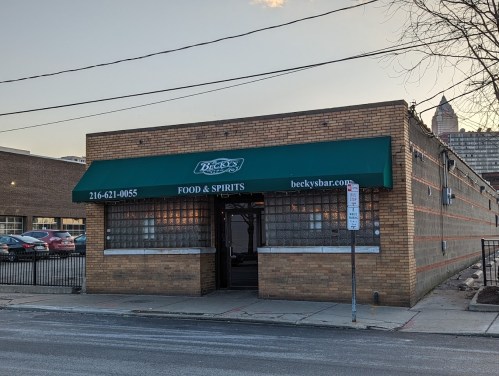 Exterior of Becky's Bar, a brick building with a green awning, featuring large windows and a sign for food and spirits.