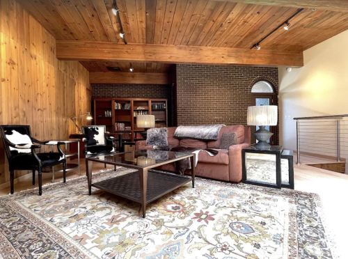 Cozy living room with wooden walls, a pink sofa, glass coffee table, and stylish lighting on a patterned rug.