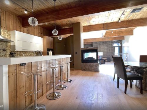 Modern kitchen with wooden beams, bar stools, and a fireplace, featuring a light-filled dining area.