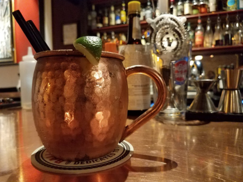A copper mug filled with a cocktail, garnished with a lime, sits on a bar counter with bottles in the background.