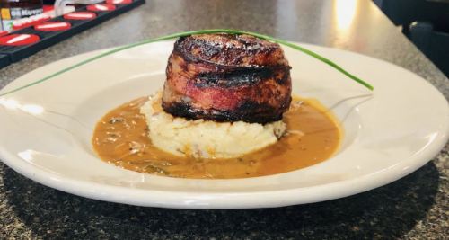 A gourmet dish featuring a bacon-wrapped meatloaf on a bed of creamy mashed potatoes with a savory sauce.