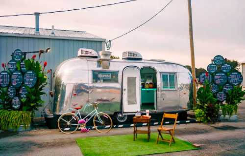 A shiny silver Airstream trailer with an "OPEN" sign, surrounded by plants and a bicycle, set in a vibrant outdoor space.