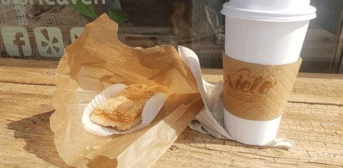 A coffee cup and a pastry on a wooden table, with a napkin and a paper wrapper nearby.