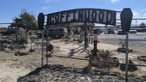 A metal gate with "Coffinwood" sign, leading to a desert area with vehicles and sparse vegetation in the background.
