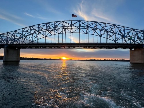 A blue metal bridge spans a river at sunset, with the sun setting behind the horizon and clouds in the sky.