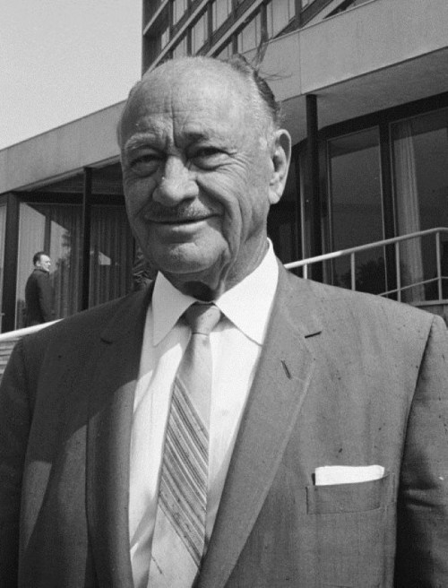 A smiling man in a suit stands outside a building, with a tie and pocket square, in a black and white photo.