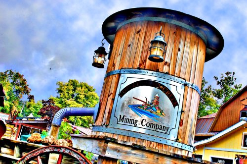 A wooden sign for "Mining Company" with lanterns, set against a cloudy sky and rustic buildings in the background.