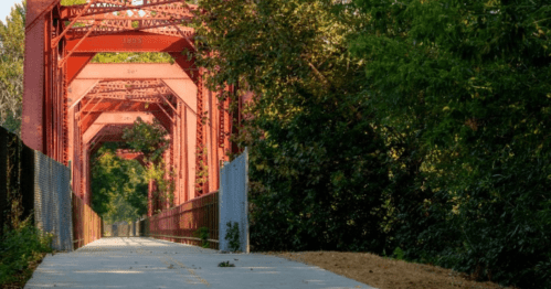 A red metal bridge spans a path surrounded by lush greenery, creating a serene outdoor scene.