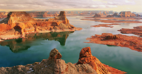 A stunning landscape of red rock formations and calm waters reflecting the sky, with distant cliffs and islands.