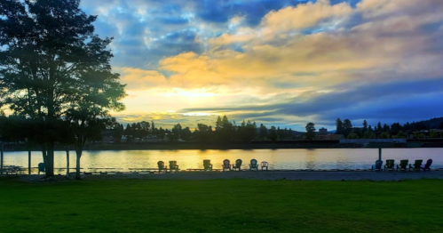 A serene lakeside view at sunset, with colorful clouds reflecting on the water and empty chairs along the shore.