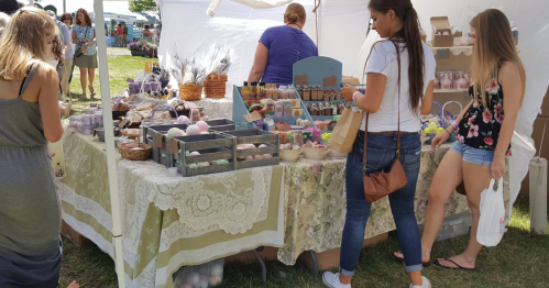 A busy outdoor market scene with people browsing a table filled with colorful handmade goods and crafts.