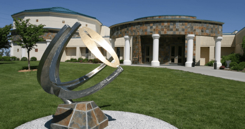 A modern sculpture in front of a stone-clad building, surrounded by green grass and trees under a clear blue sky.