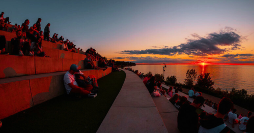People sitting on steps by a lake, watching a vibrant sunset with clouds reflecting orange and pink hues.