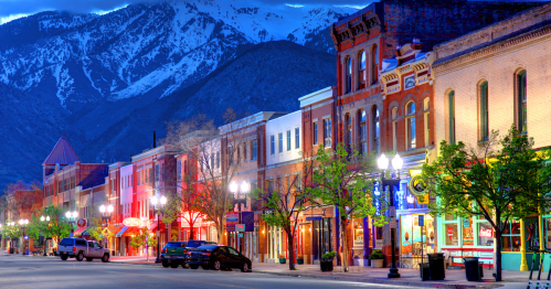 A charming street lined with historic buildings, trees, and colorful lights, set against a backdrop of mountains at dusk.