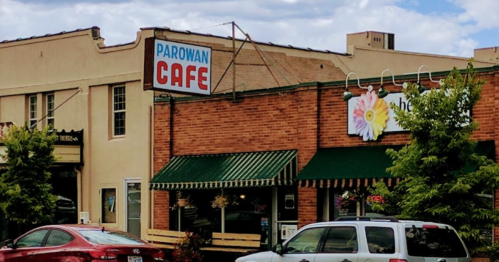 A brick building with a sign reading "Parowan Cafe" and a flower mural, featuring outdoor seating and parked cars.
