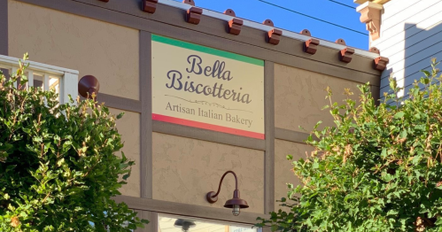 Sign for "Bella Biscotteria," an artisan Italian bakery, displayed on a building with greenery below.