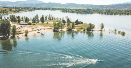 Aerial view of a serene lake with boats, sandy shore, and lush trees under a clear sky.