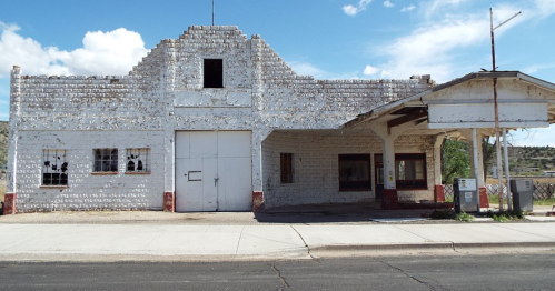 Abandoned white building with broken windows and a flat roof, set against a blue sky with scattered clouds.