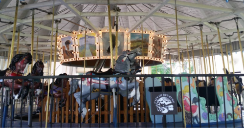 A vintage carousel with painted horses and a colorful backdrop, illuminated by lights under a wooden pavilion.