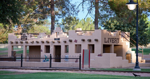 A small, adobe-style play structure resembling a western town with a bank and livery, surrounded by trees.