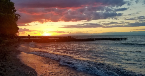 Sunset over a calm beach with a wooden pier, colorful clouds reflecting on the water's surface.