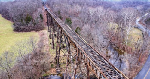 Aerial view of an old, rusty railway trestle bridge over a creek, surrounded by bare trees and grassy fields.
