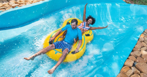 A man and woman joyfully ride a yellow inflatable tube down a water slide, splashing in the blue water.