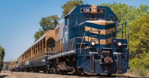 A blue and yellow locomotive pulls a vintage train past a sign for Haviland, surrounded by greenery.