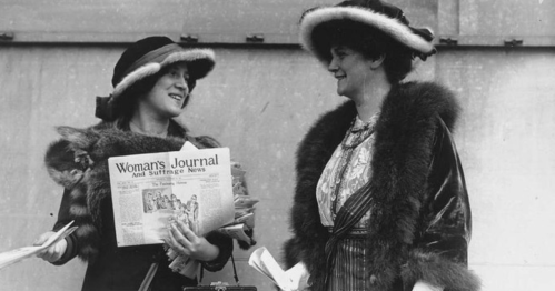 Two women in vintage hats and fur coats, smiling and holding a newspaper titled "Woman's Journal and Suffrage News."
