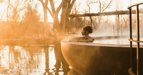 A woman relaxes in a steaming outdoor pool at sunset, surrounded by mist and trees.