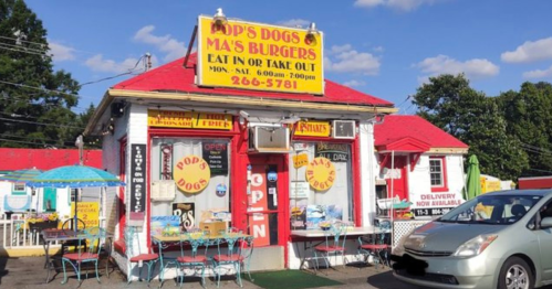 A colorful fast-food restaurant with a red roof, featuring signs for hot dogs and burgers, and outdoor seating.