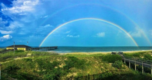 A vibrant double rainbow arcs over a beach pier, with blue skies and green dunes in the foreground.