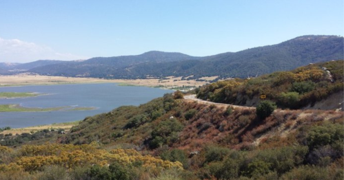 Scenic view of a lake surrounded by hills and greenery, with a winding road in the foreground under a clear blue sky.