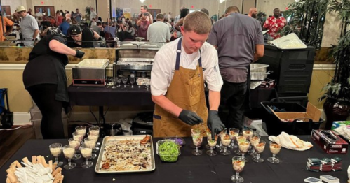 A chef prepares small dishes at a busy event, with guests in the background and a variety of food on the table.