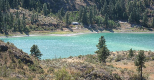 A serene lake surrounded by green trees and rocky terrain, with a small cabin visible in the distance.