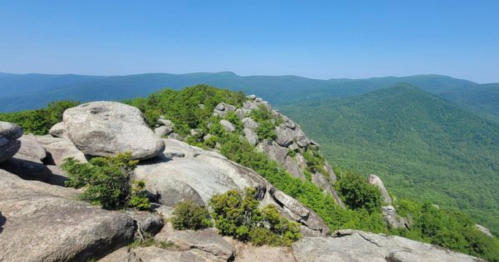 A rocky outcrop overlooks a lush green valley and distant blue mountains under a clear blue sky.