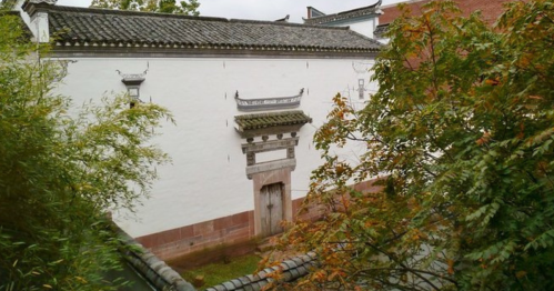A traditional white building with a wooden door, surrounded by greenery and a tiled roof in the foreground.