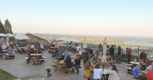 A lively outdoor dining area with people socializing, tables, and umbrellas, overlooking a scenic landscape.