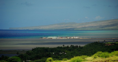 A scenic view of a coastal area with a small harbor, lush greenery, and distant mountains under a blue sky.