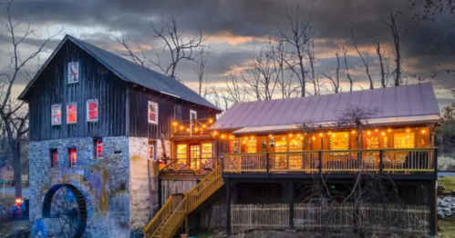 A rustic building with a water wheel, adorned with warm lights, set against a dramatic sky at dusk.