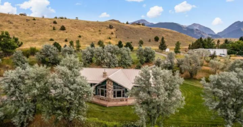 A scenic view of a house surrounded by trees, with rolling hills and mountains in the background under a blue sky.