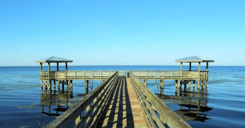 A wooden pier extends over calm water, leading to two gazebos under a clear blue sky.