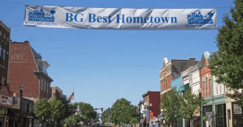 A banner reading "BG Best Hometown" hangs over a sunny street lined with shops and trees in Bowling Green.