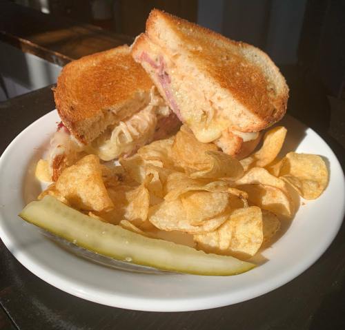A toasted sandwich with melted cheese and ham, served with potato chips and a pickle on a white plate.