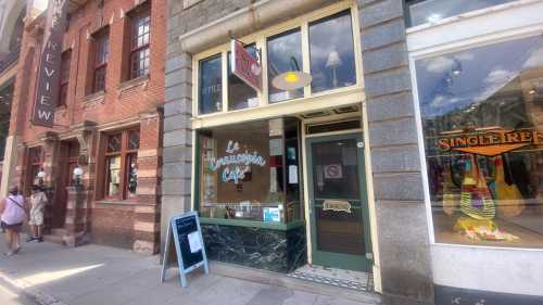 Exterior of Le Trapezium Café, featuring large windows and a sign, with pedestrians walking by on a sunny day.