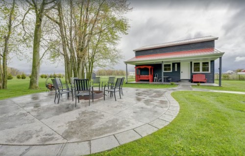 A cozy house with a red roof, surrounded by green grass and trees, features a circular patio with a fire pit and chairs.