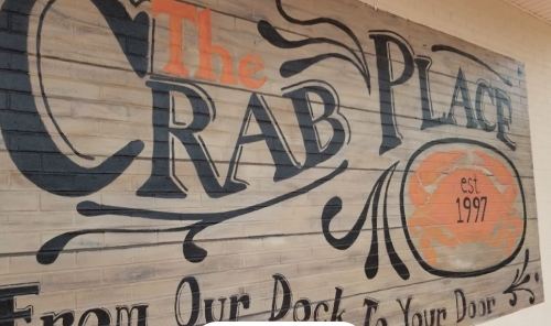 Sign for "The Crab Place," established in 1997, featuring decorative lettering and a tagline about fresh seafood delivery.