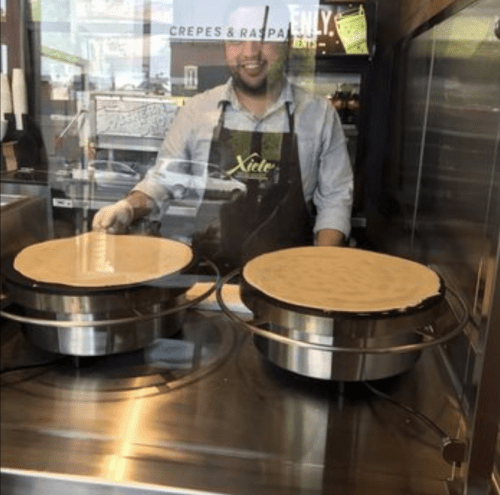 A smiling man in an apron stands behind a counter with two crepe makers, preparing fresh crepes.